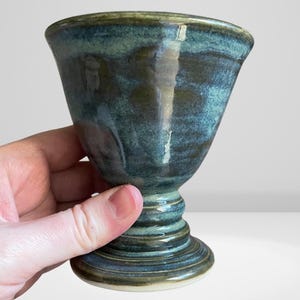 Könnte beinhalten: Ein handgefertigter Keramikbecher mit ausgestelltem Rand und Sockel. Der Becher ist in Blau- und Grüntönen glasiert und hat ein strukturiertes Aussehen. Der Sockel hat eine Reihe konzentrischer Ringe.