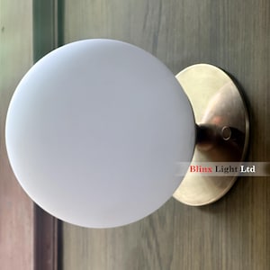 May include: A wall-mounted light fixture featuring a frosted white globe shade and a circular brass-colored base. The fixture is mounted on a wooden surface. The text "Blinx Light Ltd" is visible.