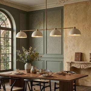 May include: A linear pendant light fitting featuring four matte beige dome-shaped shades suspended from a thin brass-toned horizontal bar. The fitting provides downward illumination over a dining table set with ceramic tableware and a vase.