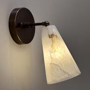 Op de afbeelding: Een wandlamp met een ronde bronzen basis en een conische, crèmekleurige kap. De kap heeft een gemarmerd uiterlijk met bruine aderen. De lamp is ontworpen om licht naar beneden te richten.