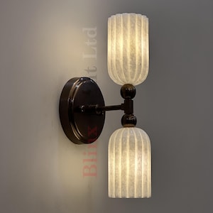 Puede incluir: Aplique de pared con acabado en bronce oscuro. Cuenta con dos pantallas de vidrio blanco roto con estrías verticales. Las pantallas están conectadas por un brazo curvo y una bola decorativa. El aplique está montado sobre una placa trasera circular.