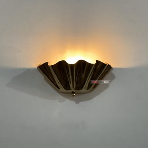 Puede incluir: Aplique de pared decorativo con un diseño festoneado en forma de abanico. El aplique está hecho de un material metálico oscuro y emite una luz cálida. El accesorio está montado en una pared gris claro.