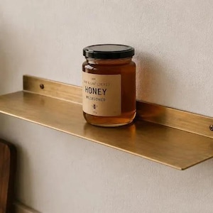 Könnte beinhalten: Ein rechteckiges Wandregal aus Messing mit gebürsteter Oberfläche und zwei sichtbaren Befestigungsschrauben. Ein Glas mit Wildblumenhonig, schwarzem Deckel und beigem Etikett mit der Aufschrift "Raw & Unfiltered Honey Wildflower" steht mittig auf der Regalfläche.