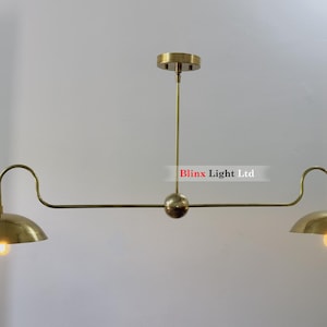 Puede incluir: Lámpara colgante de latón con dos luces. El accesorio tiene un vástago central y dos brazos curvos, cada uno con una pantalla en forma de cúpula. Las bombillas están encendidas. El texto "Blinx Light Ltd" es visible. Adecuado para interiores modernos.