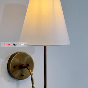 Puede incluir: Aplique de pared con pantalla blanca y base de metal color latón. La pantalla tiene forma de cono y el brazo metálico es curvo. El aplique está diseñado para montarse en la pared y proporcionar iluminación ambiental.
