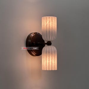 Può includere: Applique da parete con finitura bronzo scuro e due paralumi in vetro bianco a coste. I paralumi sono cilindrici ed emettono una calda luce. L'applique è montata su una piastra rotonda. Il testo "Bliss Light Ltd" è visibile.