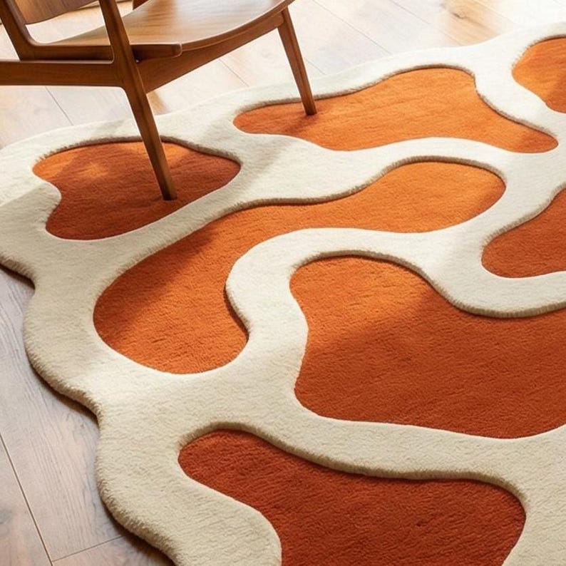 May include: An orange and cream-colored rug with an abstract design. The rug features a series of interconnected, organic shapes in a warm orange hue, outlined by a cream-colored border. The rug is made of a soft, plush material.