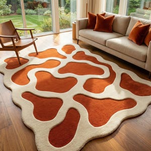 May include: An abstract area rug with a unique design. The rug features a burnt orange color with cream-colored, amoeba-shaped patterns. The rug is placed on a hardwood floor in a living room setting, with a sofa and chair visible.