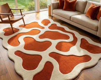 May include: An abstract area rug with a unique design. The rug features a burnt orange color with cream-colored, amoeba-shaped patterns. The rug is placed on a hardwood floor in a living room setting, with a sofa and chair visible.
