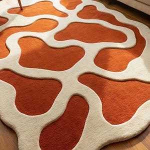 May include: A round rug with an abstract design. The rug features a pattern of orange shapes outlined in off-white. The rug is made of a soft, textured material and is placed on a wooden floor. The rug's design adds a modern touch to any room.