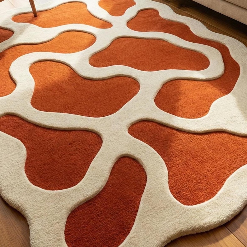 May include: A round rug with an abstract design. The rug features a pattern of orange shapes outlined in off-white. The rug is made of a soft, textured material and is placed on a wooden floor. The rug's design adds a modern touch to any room.