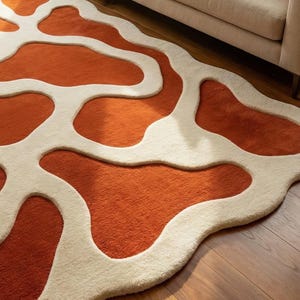 May include: An orange and cream-colored area rug with an abstract, organic pattern. The rug features a plush, textured surface with a design of flowing, irregular shapes in a contrasting cream color against the orange background. The rug is placed on a wooden floor.