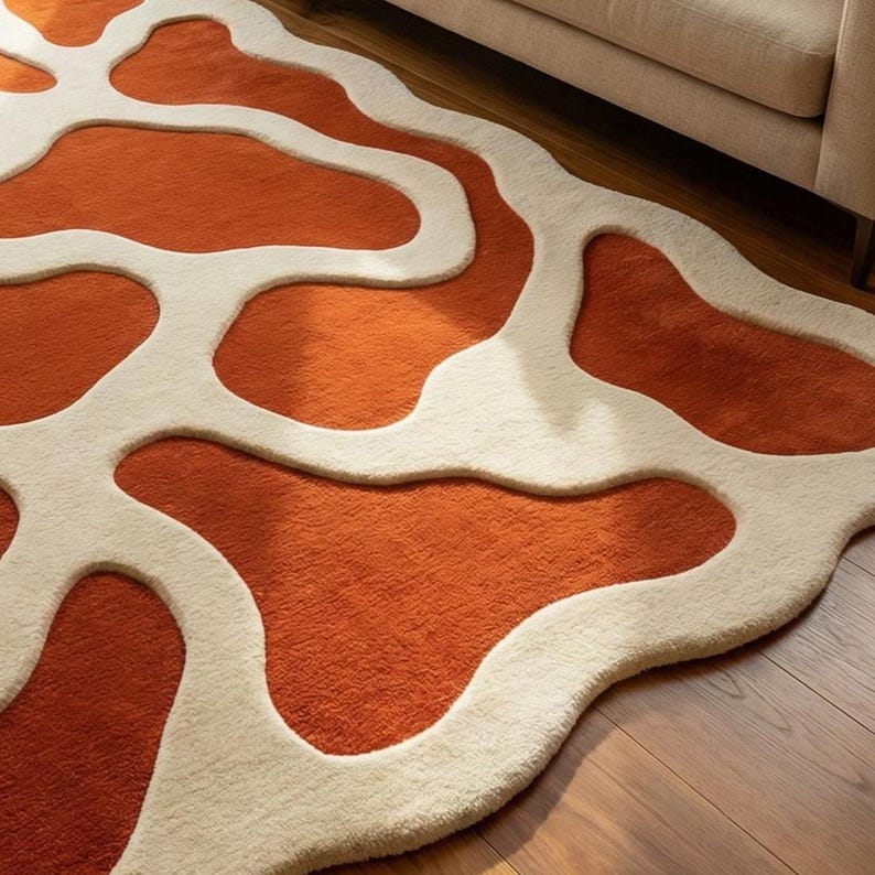 May include: An orange and cream-colored area rug with an abstract, organic pattern. The rug features a plush, textured surface with a design of flowing, irregular shapes in a contrasting cream color against the orange background. The rug is placed on a wooden floor.