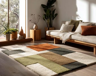 May include: A rectangular area rug with a geometric pattern in shades of orange, green, brown, and cream. The rug is placed on a hardwood floor in a well-lit room, with a sofa and plants in the background.