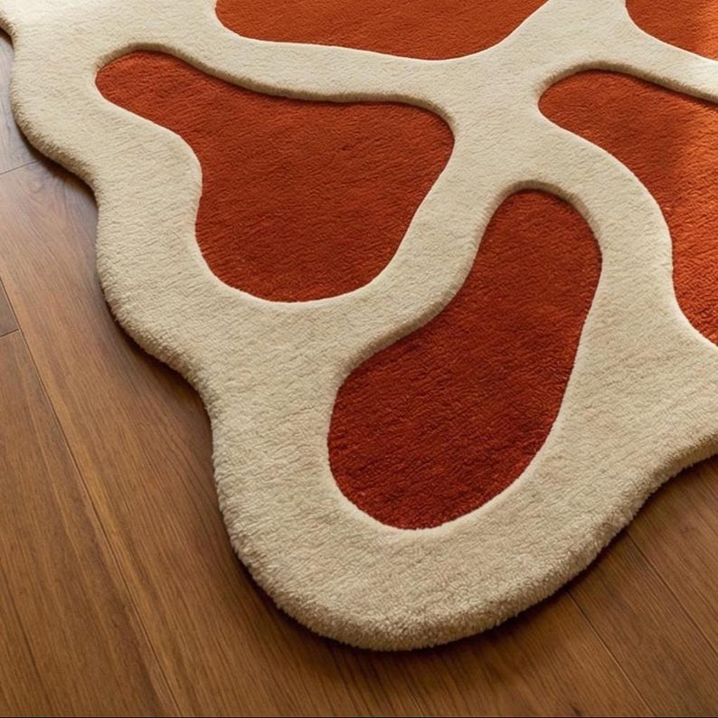 May include: A modern area rug with an abstract design. The rug features a burnt orange background with an ivory-colored, amoeba-like pattern. The rug is made of a soft, textured material and is placed on a wooden floor.