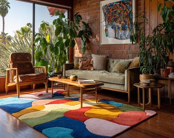 May include: A colorful area rug with an abstract design dominates the foreground of a living room. The rug features large, irregular shapes in shades of blue, red, yellow, and green. The room includes a green sofa, wooden furniture, and a large window with a view.