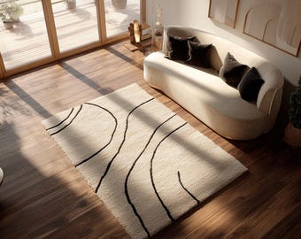 May include: A rectangular, cream-colored rug with a black, abstract, wavy line design. The rug is placed on a dark wood floor in a living room setting. A curved beige sofa with black pillows is in the background.