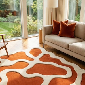May include: An orange and cream-colored area rug with an abstract design. The rug is in a living room setting, with a beige sofa and rust-colored throw pillows. Large windows offer a view of a green lawn and trees.