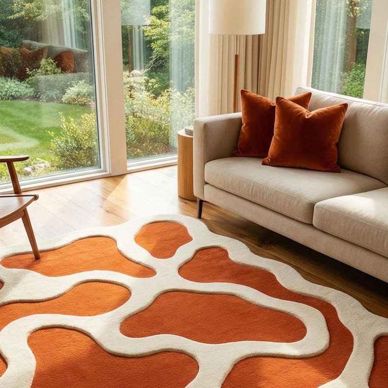 May include: An orange and cream-colored area rug with an abstract design. The rug is in a living room setting, with a beige sofa and rust-colored throw pillows. Large windows offer a view of a green lawn and trees.
