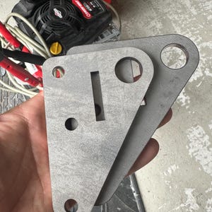 May include: Two gray metal brackets with various holes and a rectangular slot. The brackets are held in a hand, showing their triangular and rectangular shapes. The metal has a rough, unfinished texture.