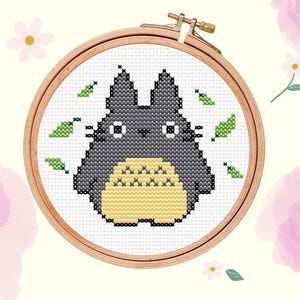 May include: Cross-stitch embroidery of a gray Totoro character with a yellow belly, surrounded by green leaves, set in a wooden hoop. The background features pink watercolor flowers and the text "Loopie Daisies".
