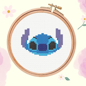 Può includere: Ricamo a punto croce del personaggio Stitch di Lilo & Stitch, nei toni del blu e del nero, inserito in un telaio da ricamo in legno. Lo sfondo è color crema chiaro con fiori rosa ad acquerello e il testo "Loopie Daisies".