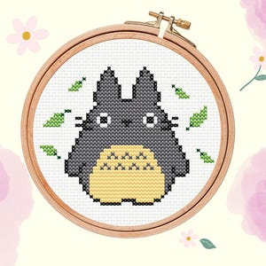 May include: A circular wooden embroidery hoop containing a cross-stitch pattern of a grey, cat-like creature with a cream-coloured belly, white eyes, and small green leaves scattered around it. The text Loopie Daisies is printed in the bottom right corner.