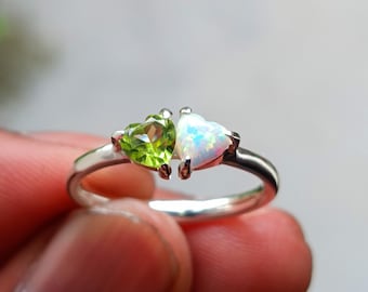 May include: A silver ring featuring two heart-shaped gemstones. One is a vibrant green, the other an iridescent white. The gemstones are set side-by-side, creating a unique and romantic design. The ring is a delicate piece of jewelry.