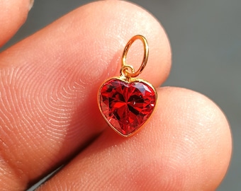 May include: A gold-colored heart-shaped pendant with a vibrant red gemstone. The pendant has a loop at the top for a chain. The gemstone is faceted, catching the light and creating a sparkling effect. Jewelry.