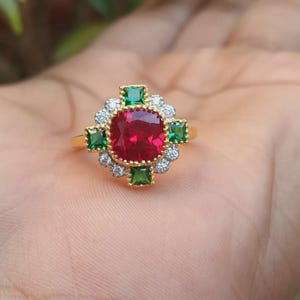 Peut inclure: Une bague en or ornée d'une grosse pierre rouge carrée au centre, entourée de petites pierres vertes et blanches. La bague présente un design détaillé et orné, de style classique et élégant.