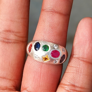 May include: A silver ring with an oval face, adorned with an array of colorful gemstones. The ring features a red, blue, green, yellow, and pink gemstone, along with several small clear stones. The ring is a statement piece.
