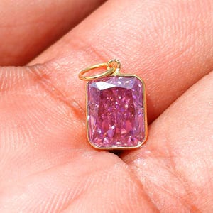 May include: A rectangular radiant-cut pink gemstone pendant set in a polished yellow gold bezel frame. The pendant features a small circular gold jump ring attached to the top, showcasing a vibrant magenta hue with multifaceted light reflections.