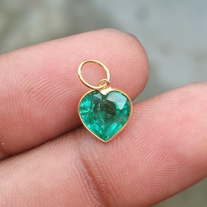 Könnte beinhalten: Ein herzförmiger Anhänger mit einem leuchtend grünen Edelstein, der in einen goldfarbenen Metallrahmen gefasst ist. Der Anhänger hat oben eine Schlaufe zum Befestigen an einer Kette oder einem Armband. Der Edelstein ist facettiert und fängt das Licht ein.