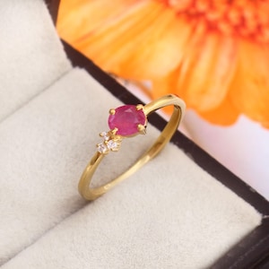 Könnte beinhalten: Ein goldener Ring mit einem runden, leuchtend roten Rubin als Hauptstein. Der Ring ist mit drei kleineren, klaren Edelsteinen verziert und vor einem weichen, neutralen Hintergrund platziert. Der Ring hat ein zartes, geschwungenes Design.