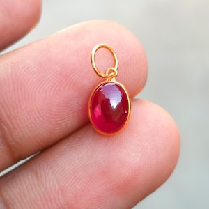 Pode incluir: Um pingente de ouro com uma vibrante pedra preciosa oval vermelha. A pedra preciosa é engastada em uma moldura de ouro e tem uma argola na parte superior para uma corrente. O pingente é segurado entre os dedos.