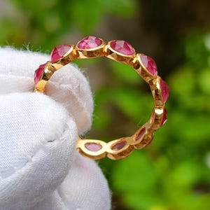 K&ouml;nnte beinhalten: Ein goldener Ring mit einer Reihe von oval geschliffenen, leuchtend rosa Edelsteinen. Die Edelsteine sind eng aneinander gesetzt und bilden ein durchgehendes Farbband. Der Ring wird vor einem verschwommenen gr&uuml;nen Hintergrund gehalten.