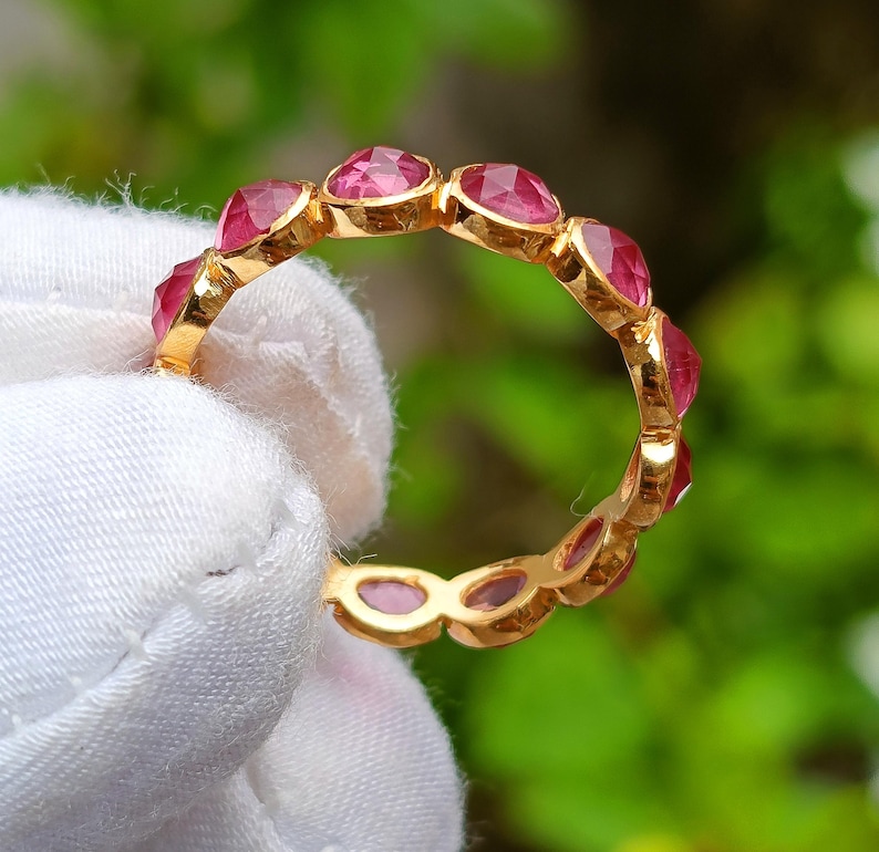 K&ouml;nnte beinhalten: Ein goldener Ring mit einer Reihe von oval geschliffenen, leuchtend rosa Edelsteinen. Die Edelsteine sind eng aneinander gesetzt und bilden ein durchgehendes Farbband. Der Ring wird vor einem verschwommenen gr&uuml;nen Hintergrund gehalten.