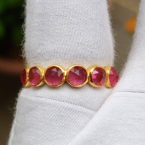 K&ouml;nnte beinhalten: Ein goldener Ring mit einer Reihe von oval geschliffenen, tiefrosa Edelsteinen. Die Edelsteine sind eng aneinander gesetzt und bilden ein durchgehendes Farbband. Der Ring wird auf einer wei&szlig;en Oberfl&auml;che gezeigt.
