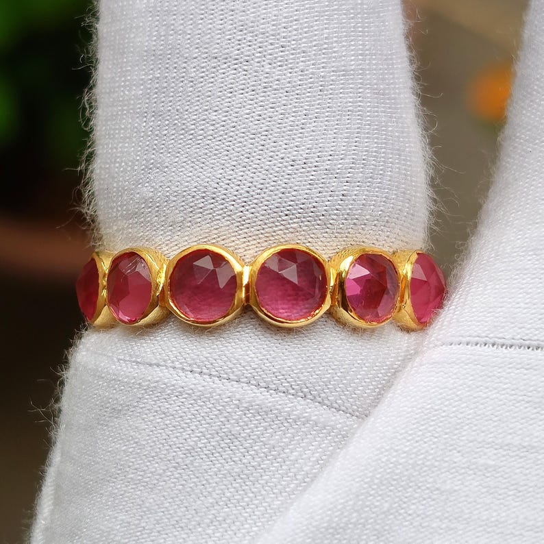 K&ouml;nnte beinhalten: Ein goldener Ring mit einer Reihe von oval geschliffenen, tiefrosa Edelsteinen. Die Edelsteine sind eng aneinander gesetzt und bilden ein durchgehendes Farbband. Der Ring wird auf einer wei&szlig;en Oberfl&auml;che gezeigt.