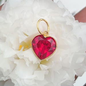 Puede incluir: Un colgante en forma de corazón con una piedra preciosa de color rojo intenso facetada y engastada en un marco de metal dorado. El colgante incluye una anilla circular dorada en la parte superior, creando un contraste con los pétalos blancos.