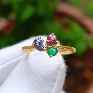 Könnte beinhalten: Ein goldener Ring mit drei Edelsteinen: einem hellvioletten Oval, einem rosa Oval und einem grünen Rund. Der Ring ist vor einem verschwommenen grünen Hintergrund und weißem Stoff platziert.