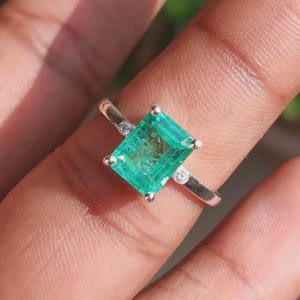 Peut inclure: Une bague en argent ornée d'une grande pierre précieuse verte de taille émeraude rectangulaire, maintenue par quatre griffes. L'anneau est agrémenté d'une petite pierre ronde transparente de chaque côté du sertissage central.
