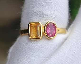 May include: A gold ring featuring two gemstones: a rectangular, yellow citrine and an oval, pink ruby. The ring's band is a thin, polished gold. The gemstones are set side-by-side, creating a unique design.