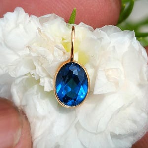 Pode incluir: Um pingente de ouro com uma pedra preciosa azul profunda, de formato oval e lapidação facetada, engastada em uma moldura de metal lisa. O pingente inclui um pequeno e fino aro de ouro no topo para fixação em uma corrente.