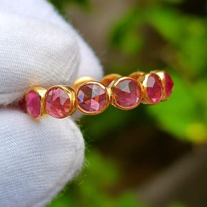 K&ouml;nnte beinhalten: Ein goldener Ring mit einer Reihe von runden, facettierten rosa Edelsteinen. Die Edelsteine sind eng aneinander gesetzt und bilden ein durchgehendes Farbband. Der Ring wird vor einem wei&szlig;en Stoffhintergrund mit verschwommenem gr&uuml;nem Hintergrund gehalten.