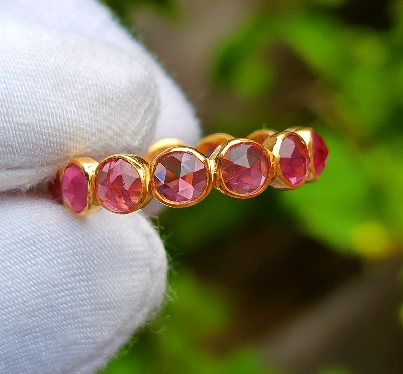 K&ouml;nnte beinhalten: Ein goldener Ring mit einer Reihe von runden, facettierten rosa Edelsteinen. Die Edelsteine sind eng aneinander gesetzt und bilden ein durchgehendes Farbband. Der Ring wird vor einem wei&szlig;en Stoffhintergrund mit verschwommenem gr&uuml;nem Hintergrund gehalten.