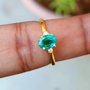 May include: A gold ring featuring an oval, emerald-colored gemstone. The ring has two small, round, clear stones on either side of the main stone. The ring is set on a gold band.