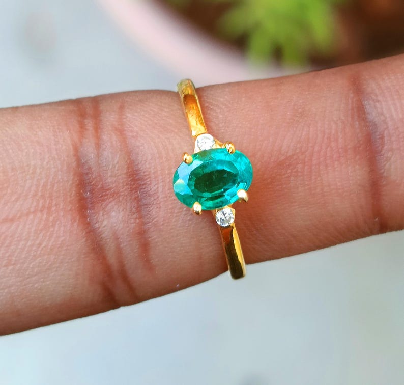 May include: A gold ring featuring an oval, emerald-colored gemstone. The ring has two small, round, clear stones on either side of the main stone. The ring is set on a gold band.