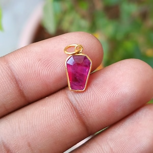 Peut inclure: Un petit pendentif doré avec une pierre précieuse rouge foncé facettée. La pierre précieuse est rectangulaire avec une forme trapézoïdale et est sertie dans une lunette dorée. Le pendentif a une petite boucle en haut pour une chaîne.