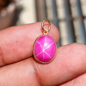 Könnte beinhalten: Ein goldener Anhänger mit einem leuchtend rosa Edelstein mit sternförmigem Muster. Der ovale Stein ist in eine goldene Lünette gefasst und hat eine Goldschlaufe für eine Kette. Der Anhänger wird von den Fingern einer Person gehalten.